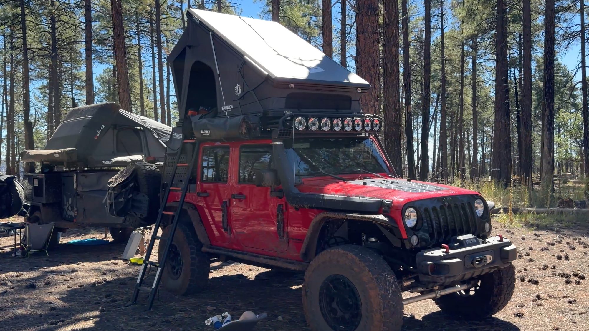 Red Jeep Wrangler with an open Taruca rooftop tent and an overland trailer camped in the pines