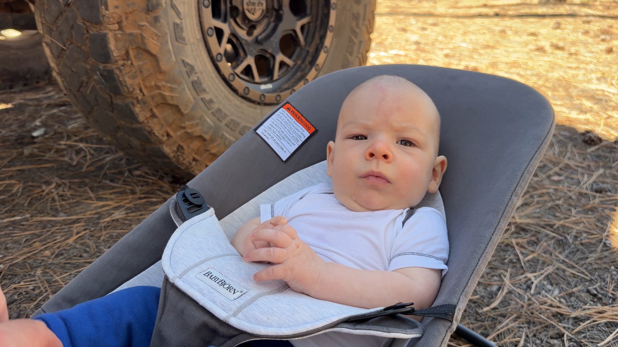 A baby in a camp bouncer seat next to a Jeep tire