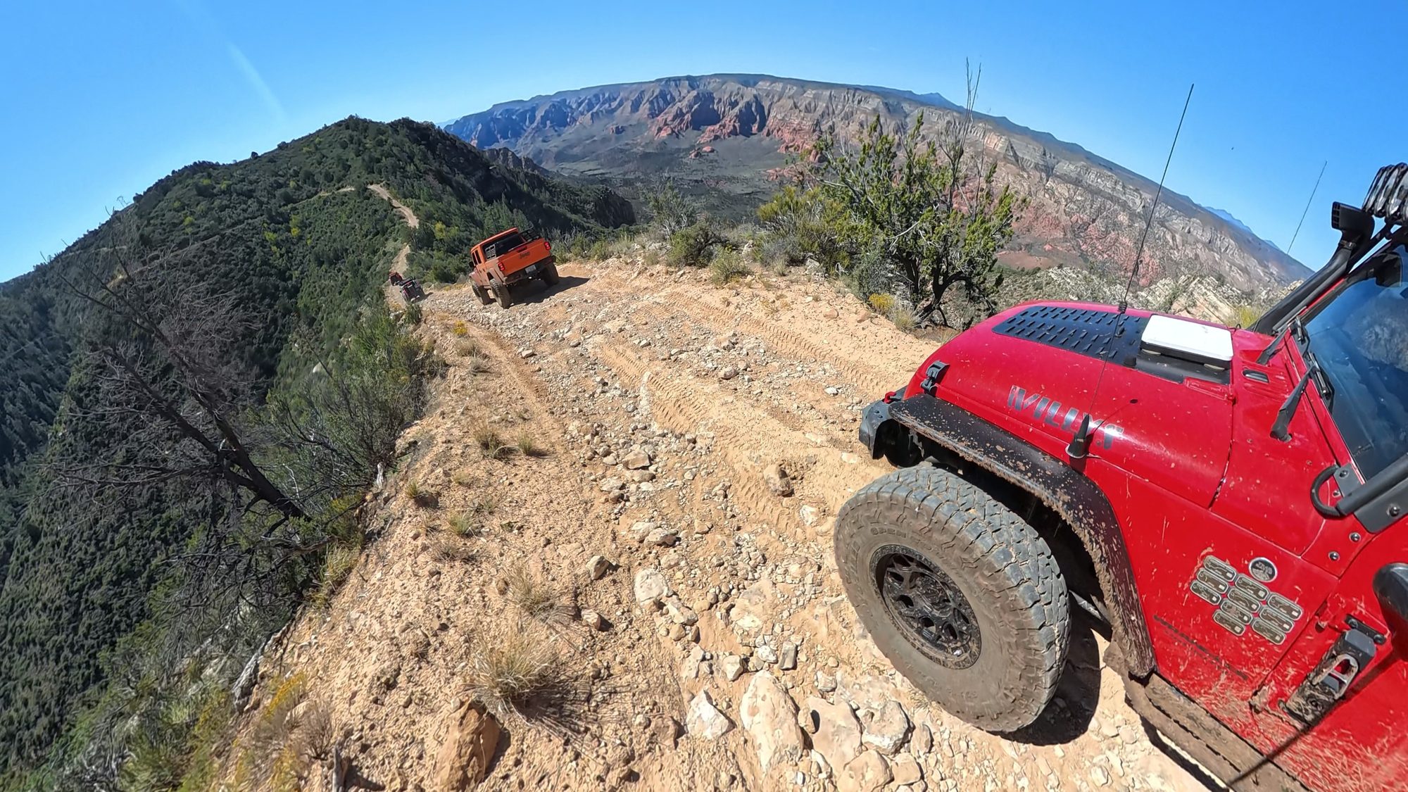 Red Jeep on a narrow ridge road with a drop-off and red rock canyon beyond