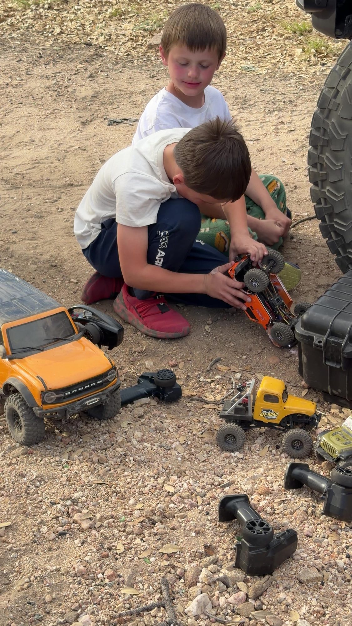 Two boys working on RC crawlers in the dirt with a lineup of scale trucks