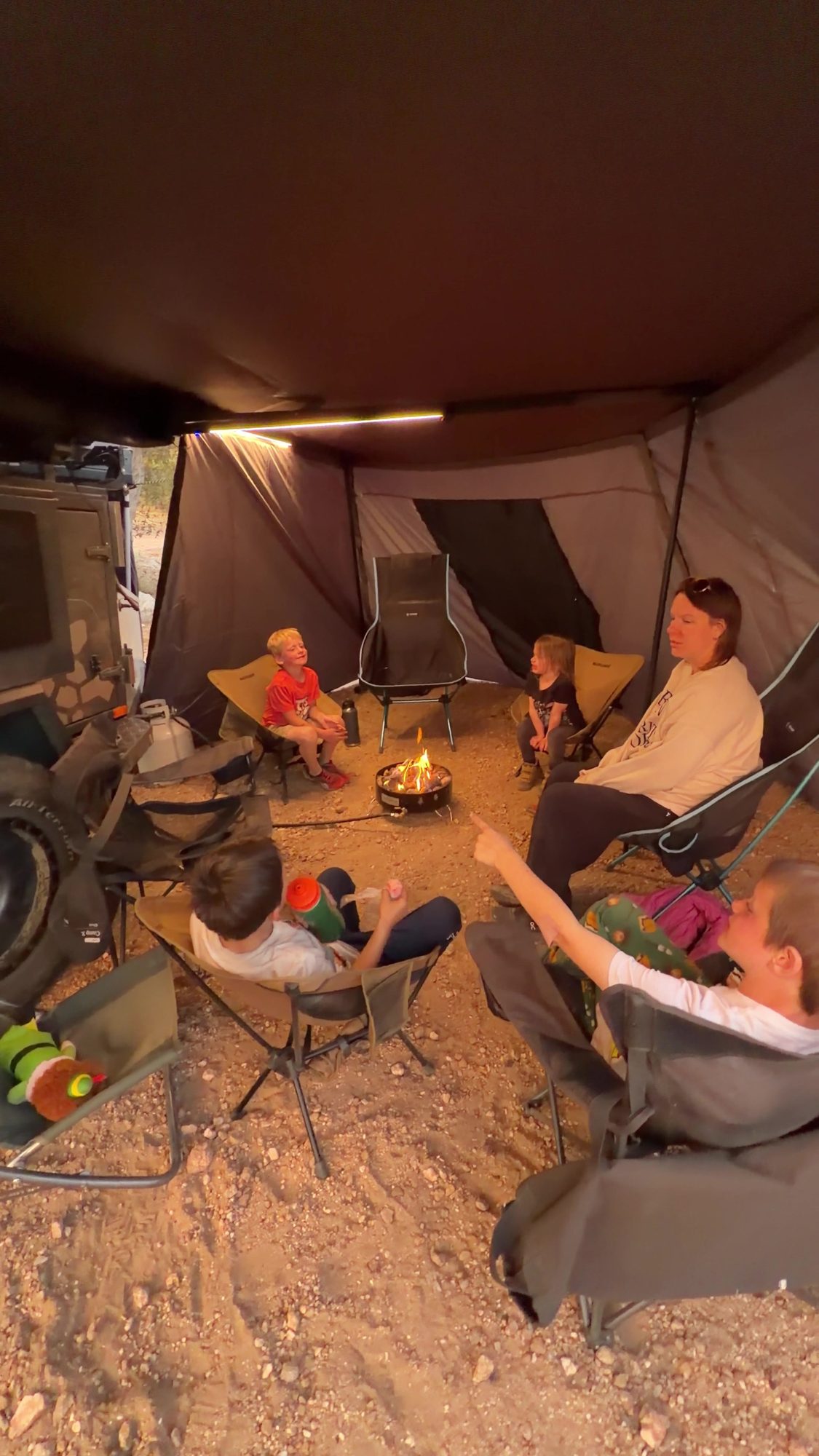 Kids and adults sitting around a propane fire pit under a Taruca awning with a wall panel blocking the wind