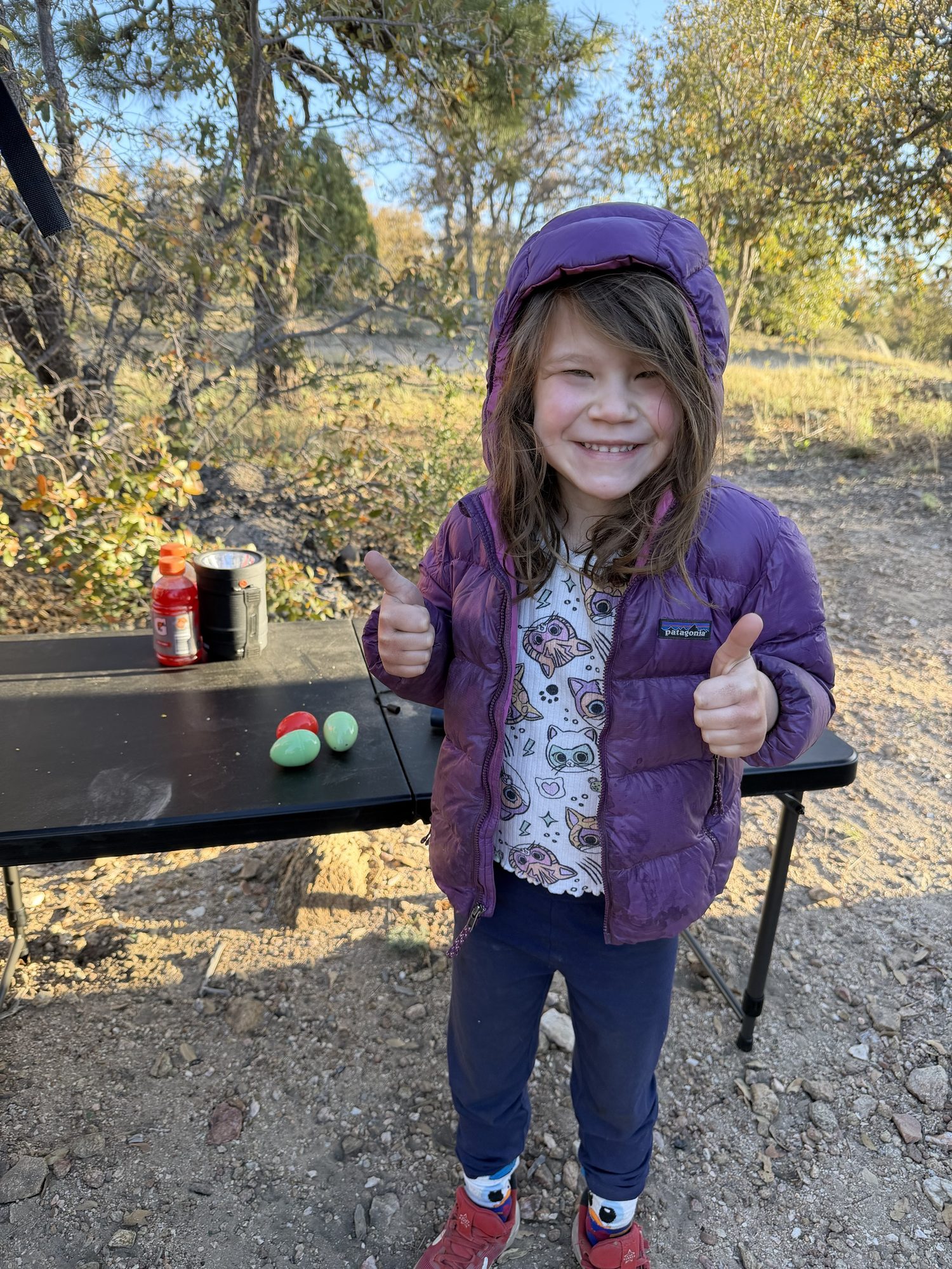 Smiling girl in a purple jacket giving two thumbs up at camp