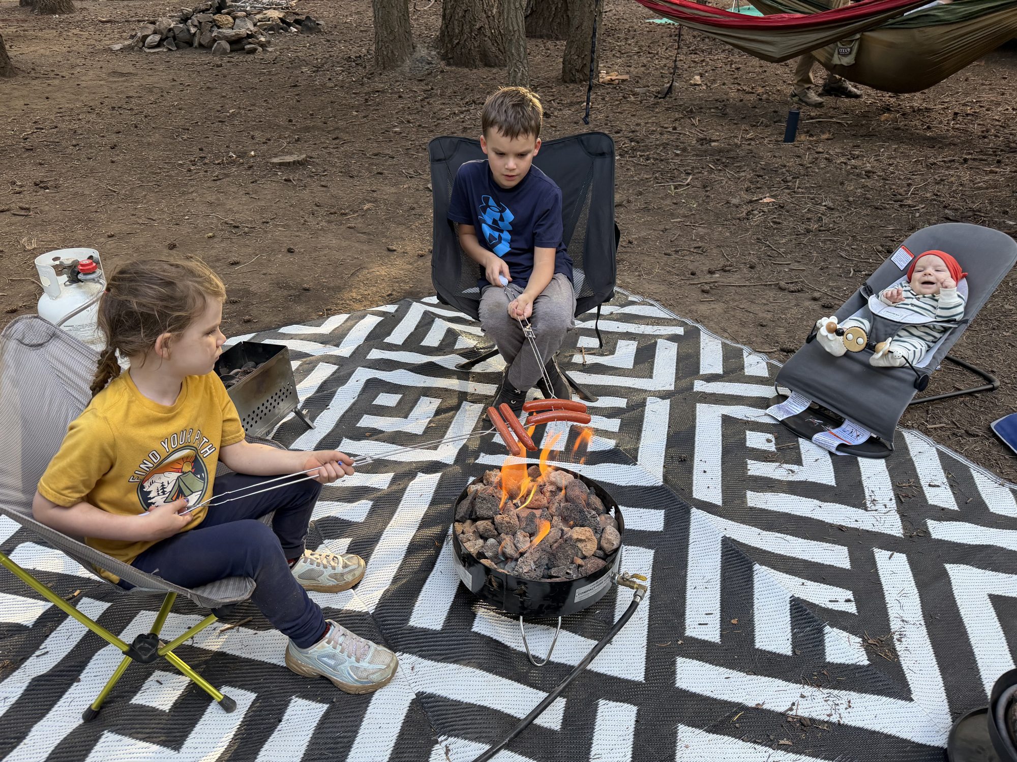 Kids roasting hot dogs over a propane fire pit at camp while a baby watches from a bouncer