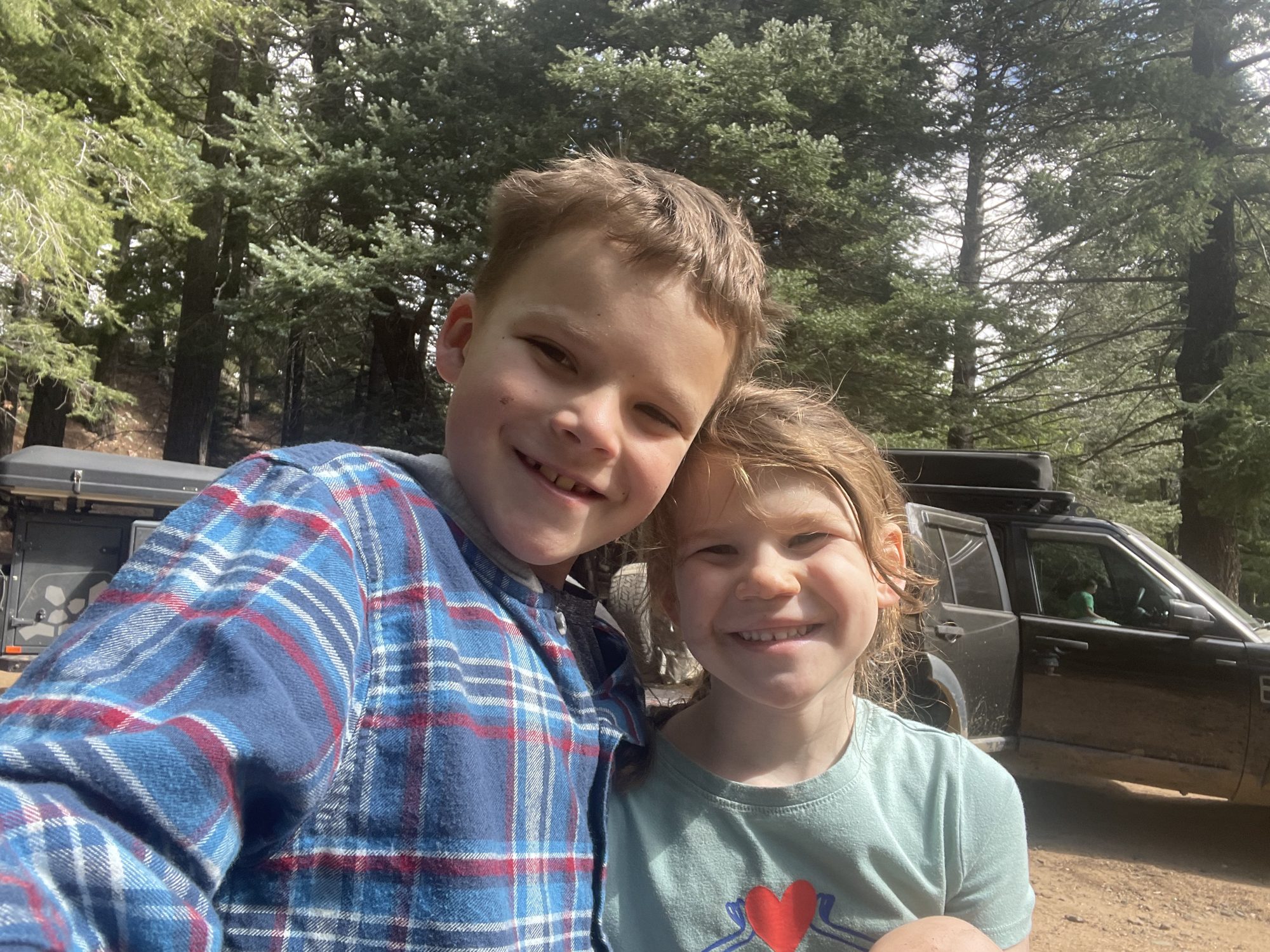Two kids smiling in front of a Land Rover LR3 and Turtleback trailer at a forest camp